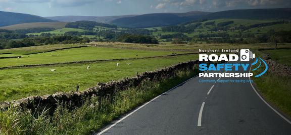 Country road surrounded by green fields with hills in the distance and logo text on right hand side that reads Northern Ireland Road Safety Partnership