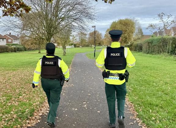 Two police officers on patrol walking along a path in a green park area.